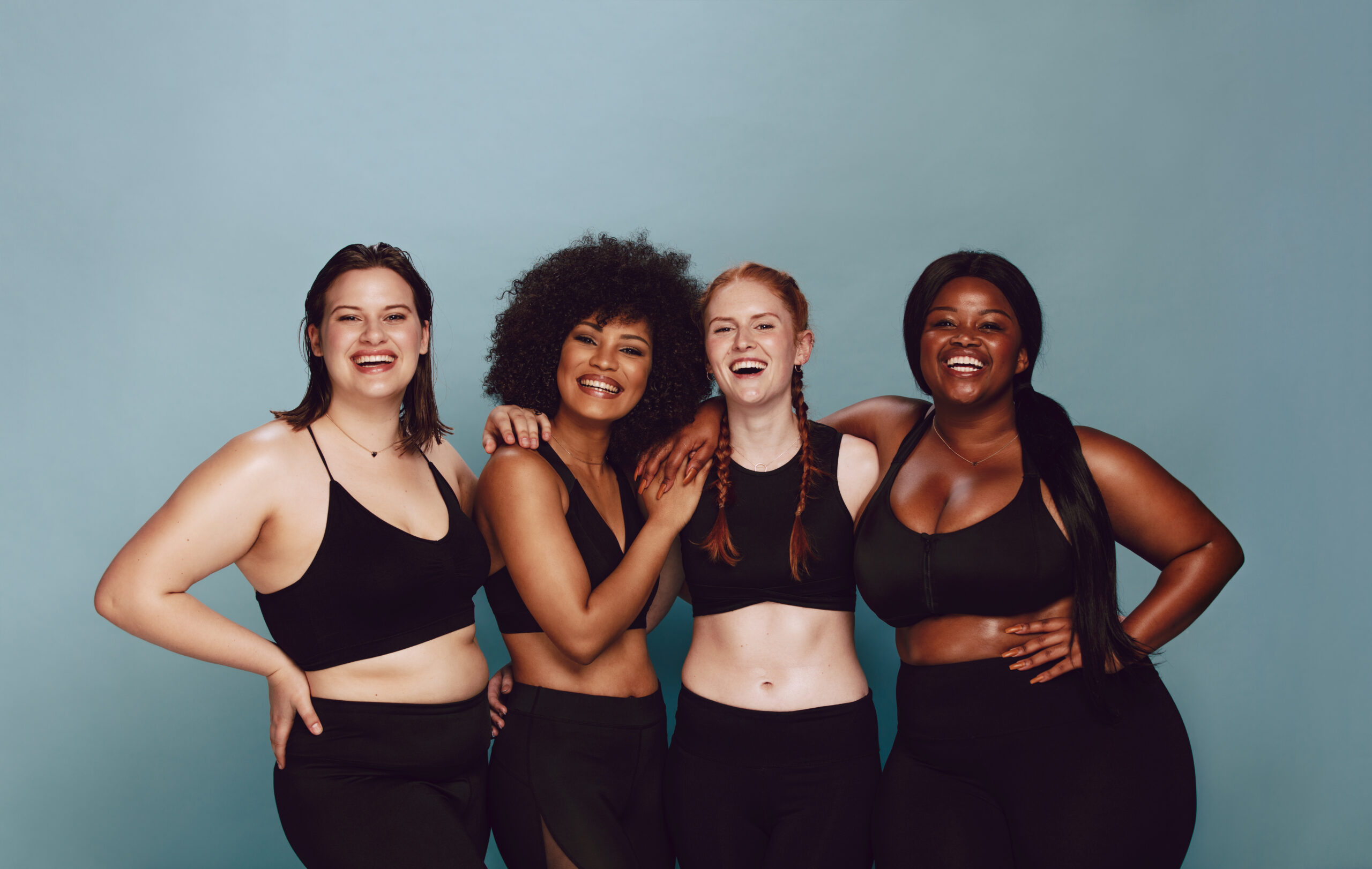 Portrait von Frauengruppen, die sich in Sportbekleidung auf grauem Hintergrund zusammensetzen. Multirassische Weibchen mit unterschiedlicher Größe stehen zusammen und schauen sich Kamera und Lächeln an.